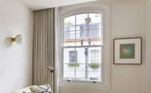 A beautifully refurbished sash window with a curved top, featuring slim-profile vacuum glazing. The window blends seamlessly into a traditional interior, offering a clear view of the period townhouses outside. The light-filled room is styled with soft neutral tones, elegant curtains, and modern decor, showcasing the balance between heritage preservation and energy efficiency.